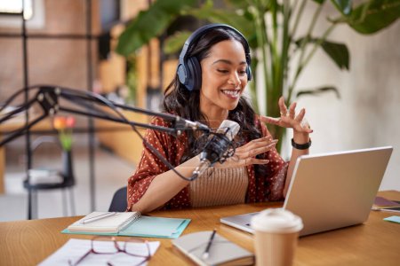 Hermoso podcast de grabación de host de radio en el estudio. Mujer joven de raza mixta grabando un podcast en el estudio. Sonriente mujer multiétnica hablando en el micrófono sobre el portátil en su estudio en casa.
