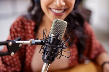 Foto de Primer plano del micrófono de pie en la mesa con podcast de grabación de mujer. Mujer latina segura grabando un podcast en el estudio usando un micrófono. Sonriente mujer multiétnica hablando sobre el detalle del micrófono. - Imagen libre de derechos