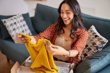 Photo for Happy young latin woman checking clothes sitting on sofa at home. Cheerful shopaholic hispanic woman holding new clothes. Smiling multuethnic girl unpacking parcel box with clothes after shopping online. - Royalty Free Image
