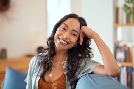 Photo for Portrait of happy young latin woman sitting on sofa at home while looking at camera. Happy multiethnic carefree woman in casual clothing relaxing at home sitting on couch. Cheerful mexican girl sitting on couch and laughing. - Royalty Free Image