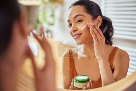 Photo for Woman caring of her beautiful skin on the face standing near mirror in the bathroom. Young mexican woman applying moisturizer on her face at home. Smiling multiethnic girl holding little green jar of bio skin cream and applying lotion on cheek. - Royalty Free Image