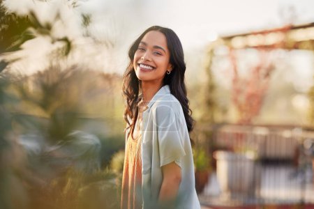 Photo for Young latin woman in casual clothing in the garden looking at camera, during early morning. Portrait of healthy mexican girl enjoying nature during sunset. Mindful multiethnic woman enjoy morning ritual with fresh air. - Royalty Free Image