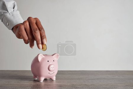Photo for Close up of black woman hand adding coin to a little pink piggy bank. African american hands of businesswoman putting money into piggy bank for saving. Home finance and clever investment strategy concept. - Royalty Free Image