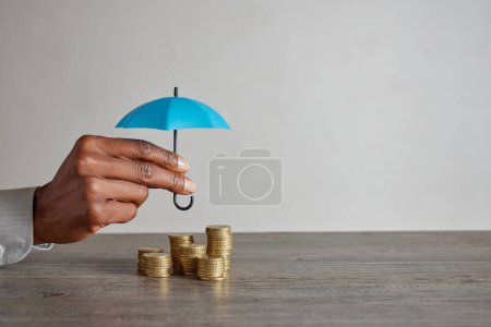 Téléchargez les photos : Gros plan d'une femme afro-américaine tenant un parapluie bleu sur une pile de pièces sur la table. Mains de conseiller financier économiser de l'argent en tenant parapluie sur les pièces. Femme d'affaires protéger l'épargne et l'argent avec l'espace de copie : sécurité financière et investissement - en image libre de droit