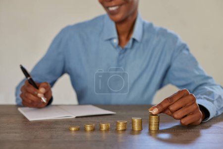 Photo for Close up of african businesswoman hands adding coins to stacks of money and writing in notebook. Smiling financial advisor managing savings after earning and noting them. Happy black woman saving money for future: finanacial investment and plan. - Royalty Free Image