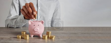 Photo for Close up of black woman hand adding money to piggy bank with stacks of coins around it. Hand of black woman putting coin to protect her saving and investment. African american girl planning her finacial future with money growth and protection, copy s - Royalty Free Image