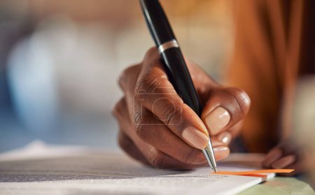 Photo for Close up of african american business woman hand writing on a contract while working in office. Successful black businesswoman signing documents using a pen while working from home. Freelancer take notes on paperwork on desk with copy space. - Royalty Free Image