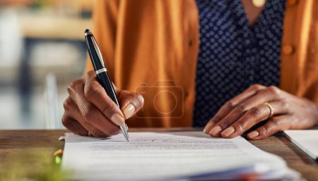 Photo for Close up of african american business woman hand writing on report while working in office. Successful black businesswoman signing documents at home using a pen. Freelancer filling contract on desk. - Royalty Free Image