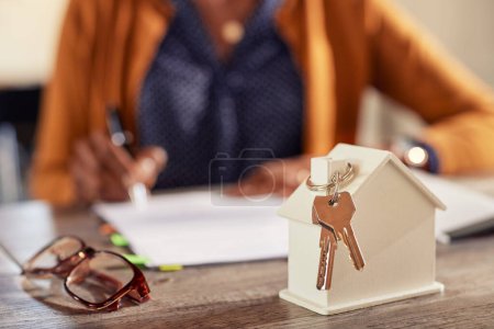 Photo for Successful independent african american woman in casual buying new home while filling out the sales documents in the background. Detail of keys from new home and tiny white model of house on work table of realtor with copy space. - Royalty Free Image