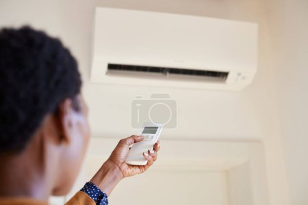 Photo for Black woman hand adjusting AC temperature with remote control to wall type air conditioning home cooling system. Hand of african woman holding remote controller directed towards the air conditioner and setting temperature. Close up of hand operating - Royalty Free Image