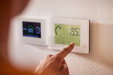 Photo for Rear view of multi ethnic woman pressing a button of a wall attached house thermostat with digital display showing the temperature. Back view of african woman adjusting digital central heating at home. Hand touching the digital thermostat at home to - Royalty Free Image