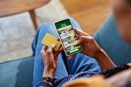 Photo for Hands of young woman using mobile phone to buy dinner from online application. Closeup of black woman order food online and pay with credit card by mobile phone. Ordering meal online and choose between sushi, pasta, salad, and burger: food delivery c - Royalty Free Image