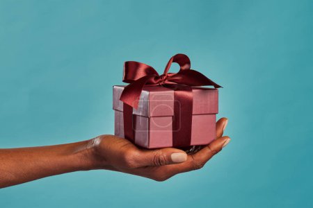 Photo for Close up shot of female hand holding a small gift wrapped with elegant brown ribbon isolated against blue background. Small craft gift in the hands of african american woman. Closeup of black woman holding elegant little box in hand for Christmas. - Royalty Free Image