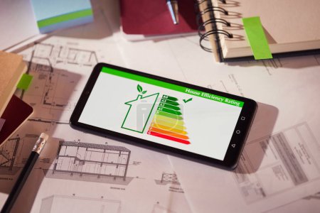 Photo for Energy efficiency screen on cellphone leaning on drawings and blueprints of a new house. Architect's desk with plans on it and a cell phone with an energy efficiency graph. Renewable energy, renovation, energy class and eco house concept. - Royalty Free Image