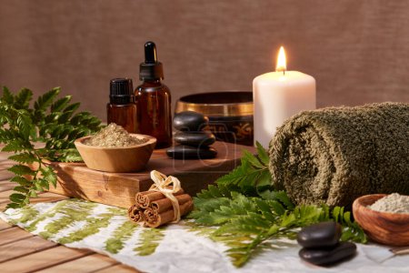Photo for Complete set of organic beauty products and accessories for natural spa treatment. Green towel on fern leaf with candle, salt and black hot stone on wooden background. Hot stone massage setting lit by candle and warm ray of light, beauty spa treatmen - Royalty Free Image