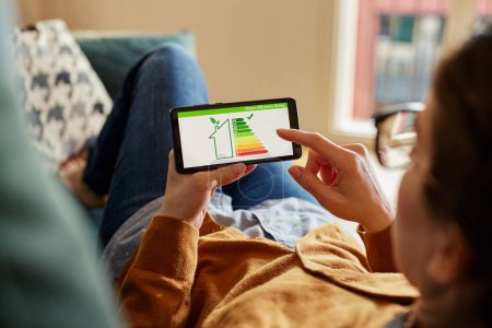 Photo for Young latin woman holding smartphone with energy efficiency chart and class on screen. Hispanic girl holding smart phone and looking at house efficiency rating. Ecological, eco renovation and bio energetic house concept. - Royalty Free Image