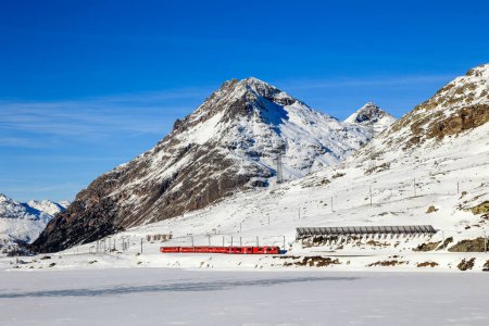 Téléchargez les photos : Berina, Switzerland - January 06. 2023: The red Rhaetian Rail is passing along the fronzen Lake Bianco with the Alps summit Piz Lagalb at the background - en image libre de droit