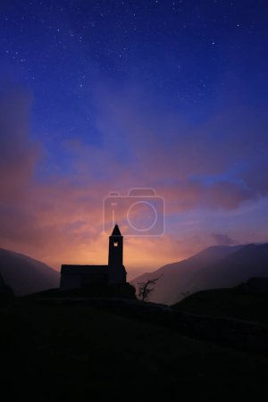 Twilight with dramatic clouds and starry sky over the chapel of San Romerio on cliff, Grisons, Switzerland