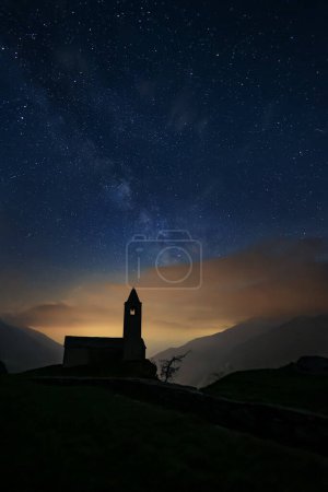 Milky Way and stars in night sky over the chapel of San Romerio on cliff, Grions, Swiss Alps