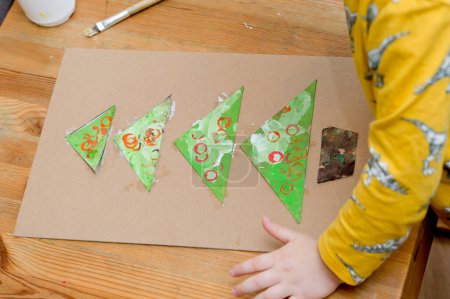 Making a Christmas tree. Boy gluing stamped colorful triangles in to the spruce shape tower. Preschool or nursery type activities at home. Montessori task for hand coordination, fosterage imagination