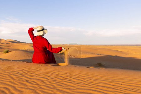 Photo for A young woman dressed in red and hat sitting on the desert dunes, playing with the sand - Royalty Free Image