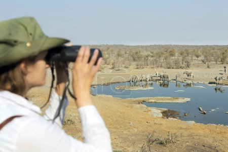 Foto de Vista trasera de una joven rubia en safari sentada en la roca mirando a través de prismáticos - concepto de viaje - Imagen libre de derechos