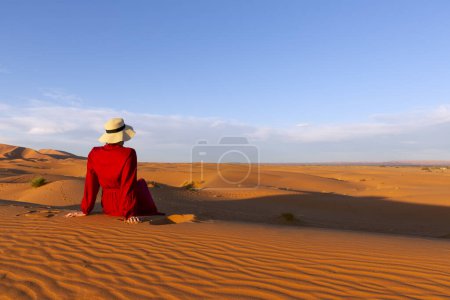 Photo for A young woman dressed in red and hat sitting on the desert dunes - Royalty Free Image