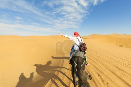 Photo for A young women  rides a camel through the dunes in the Sahara Desert. View of the woman from behind, in the background, small silhouettes of other tourists. Merzouga, Morocco - Royalty Free Image