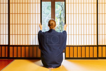 Téléchargez les photos : Maison japonaise traditionnelle ou ryokan avec femme caucasienne gaijin en kimono et tabi chaussettes ouverture shoji portes coulissantes en papier assis sur tatami tapis plancher - en image libre de droit