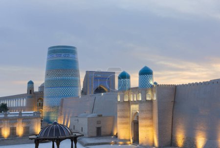 Historische Architektur des Minaretts Kalta Minor auf dem Platz und Burgtor mit Beleuchtung bei Sonnenuntergang in Itchan Kala Innenstadt der Stadt Chiwa, Usbekistan.
