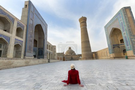Touristin mit Hut und rotem Kleid sitzt auf dem Poi Kalyon Platz, einem alten öffentlichen Platz im Herzen der antiken Stadt Buchara, Usbekistan.