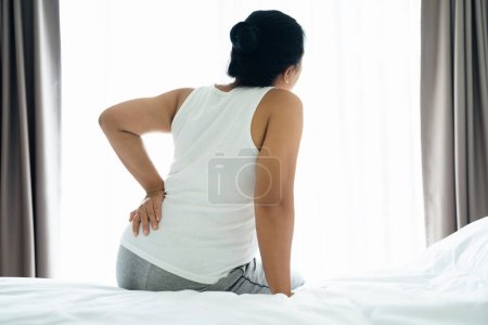 Foto de Mujer asiática adulta está sentada en la cama y sosteniendo su espalda baja sufriendo de espalda lesionada. Concepto de atención médica y dolor de espalda. - Imagen libre de derechos