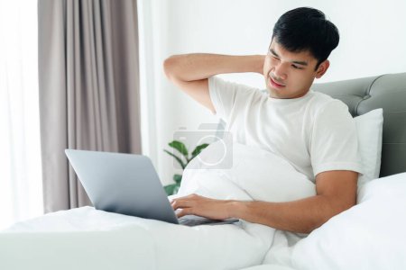 Foto de Hombre asiático en camiseta blanca acostado en la cama usando computadora portátil que tiene dolor de cuello. - Imagen libre de derechos