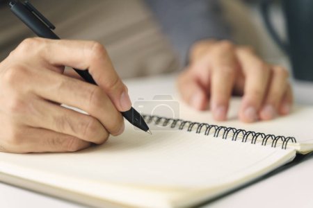 Téléchargez les photos : Gros plan de jeune homme en tissu décontracté mains écriture sur le bloc-notes, ordinateur portable à l'aide d'un stylo bille sur la table. - en image libre de droit