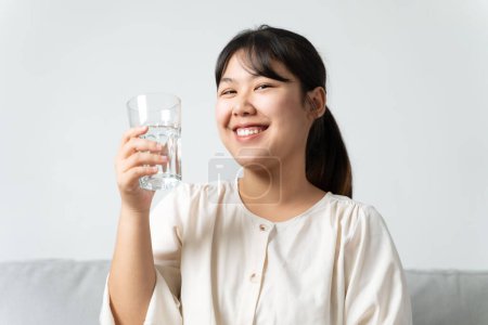 Photo for Healthy beautiful young woman holding a glass of water sitting on the couch at the livingroom. - Royalty Free Image