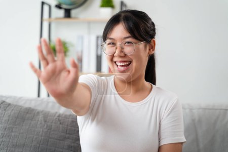Téléchargez les photos : Portrait de jeune femme asiatique portant des lunettes de vue grand sourire et regardant la caméra avec le sentiment heureux et positif dans le salon à la maison, réunion, vidéoconférence. - en image libre de droit