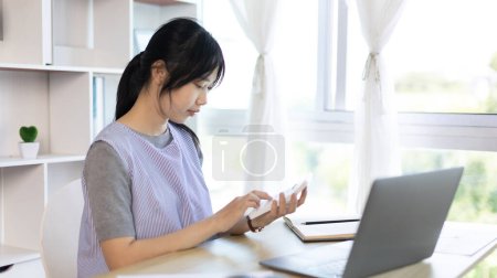 Téléchargez les photos : Femme asiatique travaillant à la maison, Appuyez sur la calculatrice pour calculer les revenus - dépenses de la maison et utiliser l'ordinateur portable pour enregistrer les informations et vérifier l'exactitude, Style de vie à la maison. - en image libre de droit
