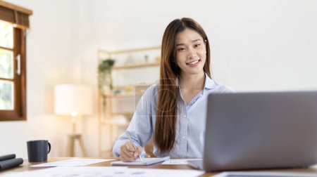 Photo for Beautiful young asian woman watching live video or video call of teacher teaching on laptop in her home, Take notes of important conversations and messages during the teacher's teaching. - Royalty Free Image