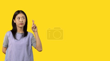 Foto de Young Asian woman making a symbolic gesture with fingers crossed showing good luck, White lie gesture, Fingers crossed, Woman doing hand sign on yellow background, Superstitious concept. - Imagen libre de derechos