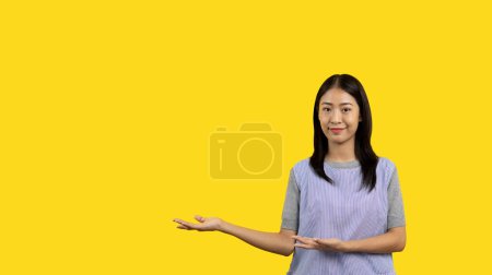 Téléchargez les photos : Asian woman with holding copyspace imaginary on the palm to insert an ad, Showing copyspace pointing, Showing her hand to present something on yellow background. - en image libre de droit