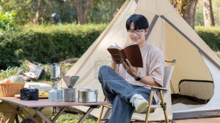 Foto de Young man camping for the weekend in the woods near the river, Camping Holiday In Countryside, Man sitting and relaxing happily reading definitions, Young man outdoor leisure activities. - Imagen libre de derechos