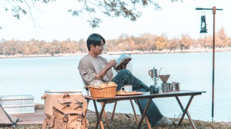 Foto de Young man camping for the weekend in the woods near the river, Camping Holiday In Countryside, Man sitting and relaxing happily reading definitions, Young man outdoor leisure activities. - Imagen libre de derechos