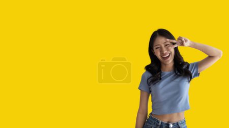 Téléchargez les photos : Young Asian woman making two thumbs up gesture showing joy and fun, Symbol of good friendship, Popular photo poses, Make a v-shaped finger, Yellow background. - en image libre de droit
