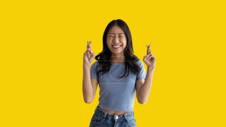 Foto de Young Asian woman making a symbolic gesture with fingers crossed showing good luck, White lie gesture, Fingers crossed, Woman doing hand sign on yellow background, Superstitious concept. - Imagen libre de derechos