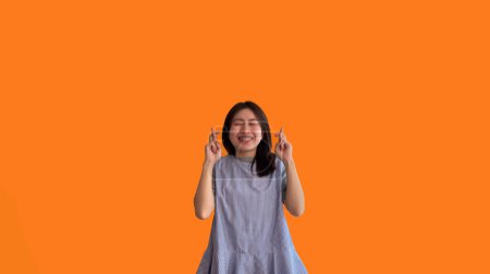 Foto de Mujer asiática joven haciendo un gesto simbólico con los dedos cruzados mostrando buena suerte, Mentira blanca gesto, Dedos cruzados, Mujer haciendo signo de mano sobre fondo naranja, Concepto supersticioso. - Imagen libre de derechos