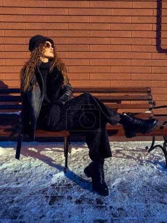 Foto de La dama del glamour se sienta en el banco al aire libre. Modelo de moda posando outddors en invierno. - Imagen libre de derechos