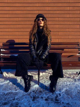 Foto de Atractiva dama se sienta en el banco al aire libre. Modelo de moda posando outddors en invierno. - Imagen libre de derechos