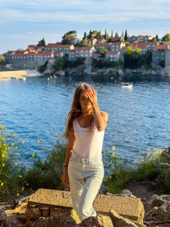 Photo for Charming woman posing at the cliff above the sea with sveti stefan on background in warm sunset lights - Royalty Free Image