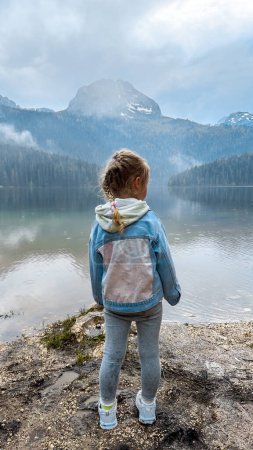 Téléchargez les photos : Fille de 3 ans regardant sur le beau lac avec des montagnes en arrière-plan au Monténégro Voyage - en image libre de droit
