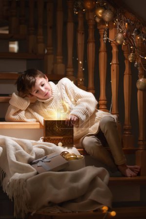Foto de Niño sentado con una fabulosa caja y regalos cerca de las escaleras en Navidad. Año Nuevo, felicidad y cuento de hadas - Imagen libre de derechos